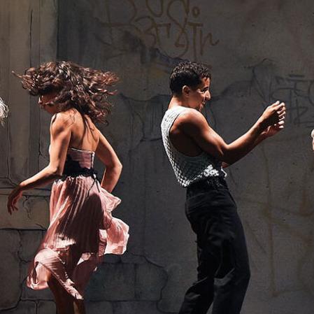A row of dancing couples on stage in front of two columns and a wall covered in graffiti